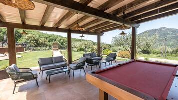 A pool table in a covered outdoor area with seating and a view of a mountainous landscape.
