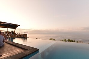 An infinity pool with a wooden deck and lounge chairs overlooking a beach. - Daphila Cottage (Penida Island)