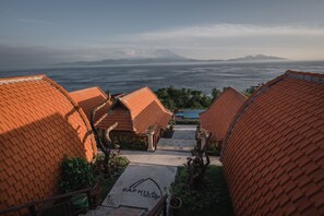 A view of Daphila Cottage with terracotta-roofed buildings, a pathway leading to the entrance, and a distant view of the sea and mountains.