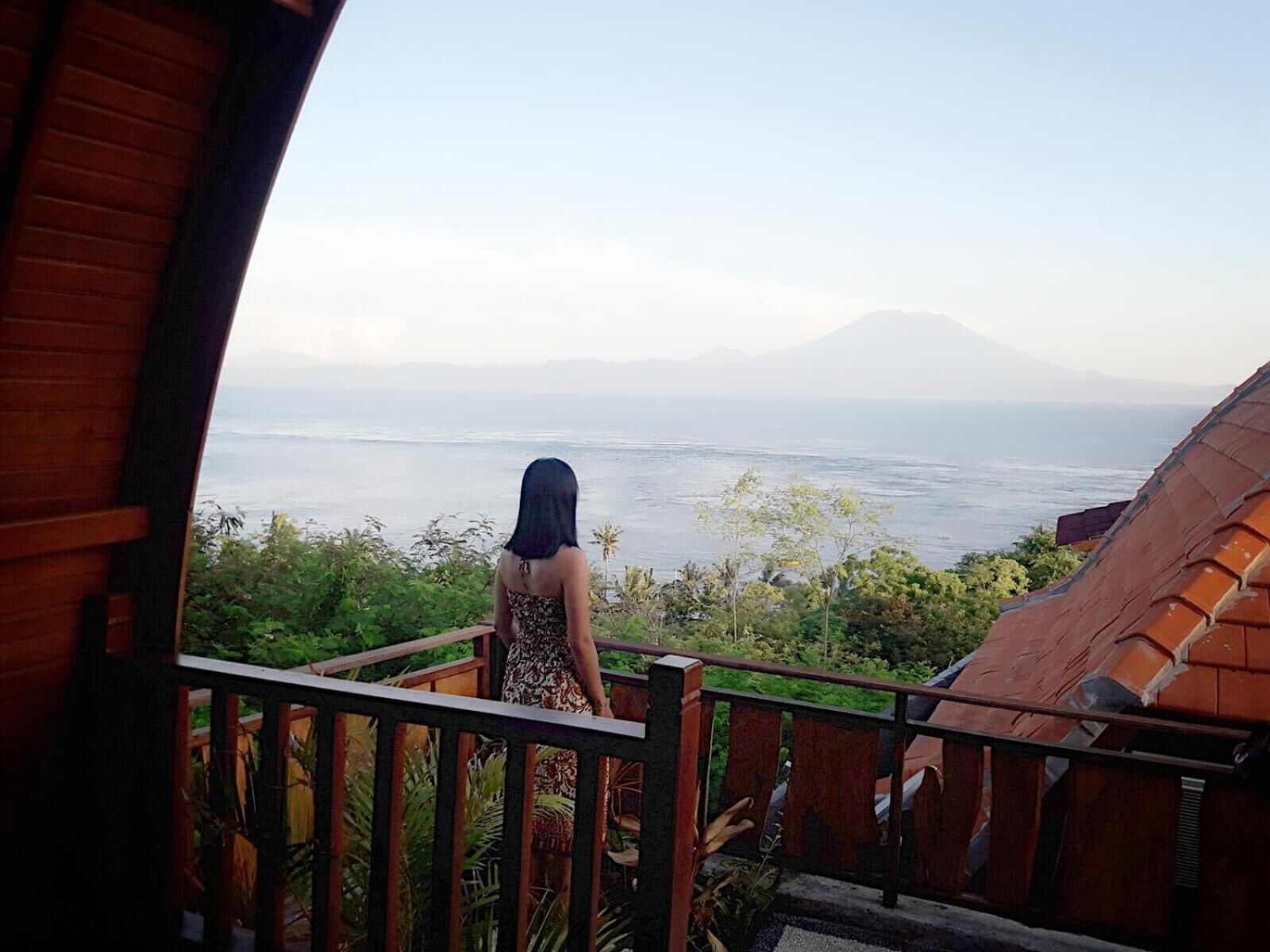 Una mujer está de pie en un balcón con vista a un cuerpo de agua y una montaña al fondo.