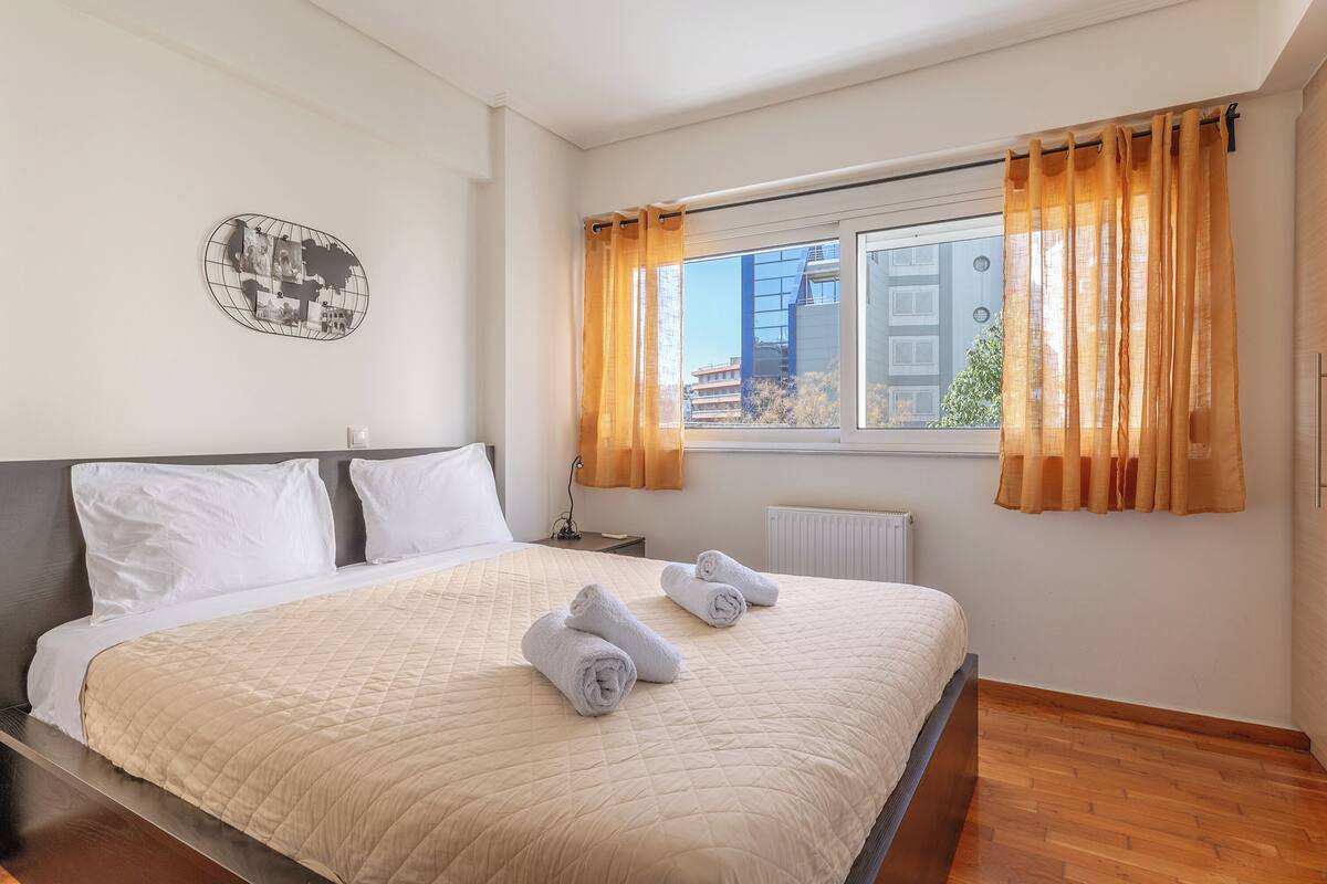 Una cama bien hecha con cubrecolcha acolchado, dos toallas enrolladas y una ventana con cortinas.