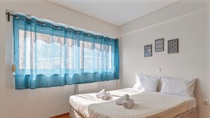 A bedroom with a bed, two pillows, a window with blue curtains, and three wall art pieces. - Elvita Apartments 2 (Athens)
