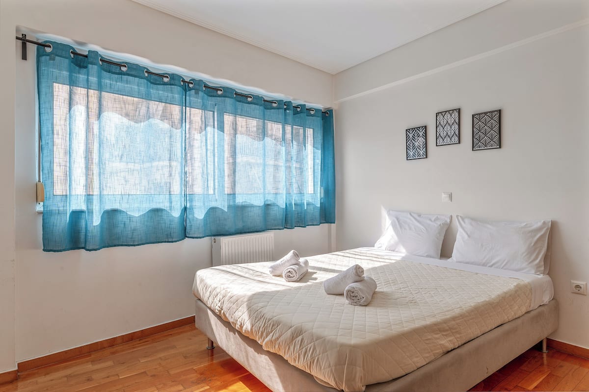 Un dormitorio con una cama, dos almohadas, un ventanal con cortinas azules y tres obras de arte en la pared.