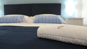 Una cama bien hecha con una colcha azul oscuro y almohadas blancas, un cabecero blanco y una mesita de noche pequeña con una lámpara.