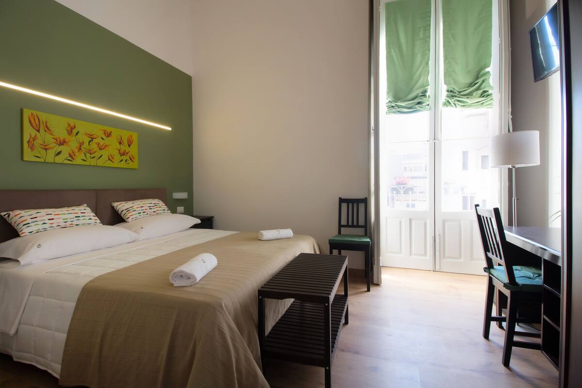 Habitación de hotel con cama, escritorio, silla, banco, un cuadro y televisor en la pared.