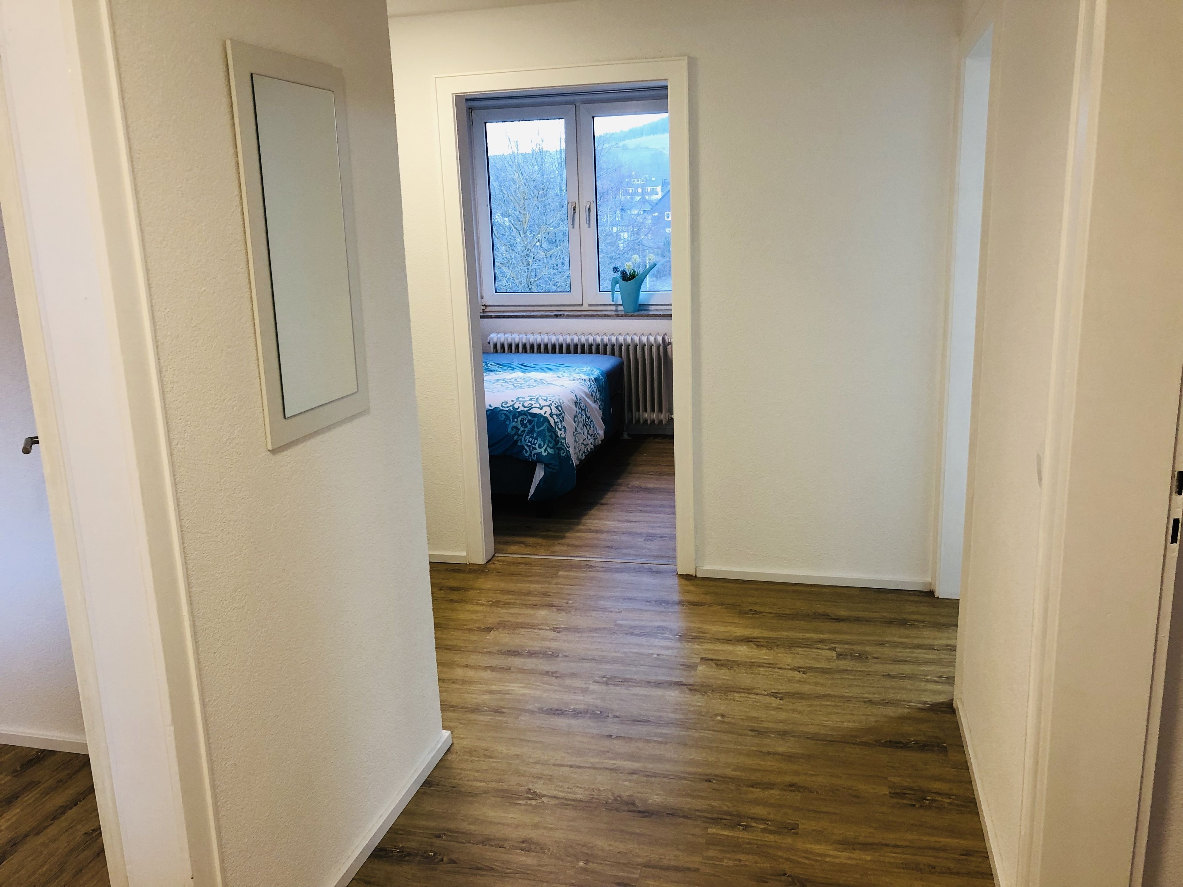 Un estrecho pasillo con piso de madera que conduce a una habitación con ventana, un radiador y una cama con colcha azul y blanca.