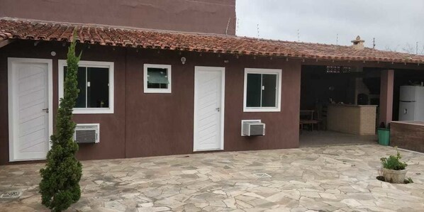A building with a tiled roof, two white doors, and a small window. There is a potted plant in the foreground and a stone-paved courtyard. - Suítes Olinda (Cabo Frio)
