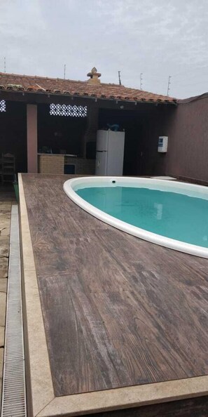 A swimming pool with a wooden deck, a tiled roof, and a refrigerator in the background. - Suítes Olinda (Cabo Frio)