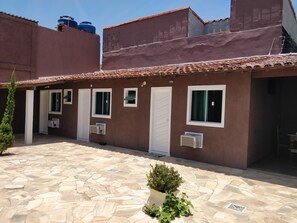 A courtyard with a stone floor, a potted plant, and a building with multiple doors and windows. - Suítes Olinda (Cabo Frio)