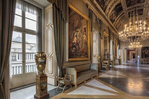 Een weelderige kamer met een imposante kroonluchter, een groot muurschildering en uitzicht op de stad door grote ramen.