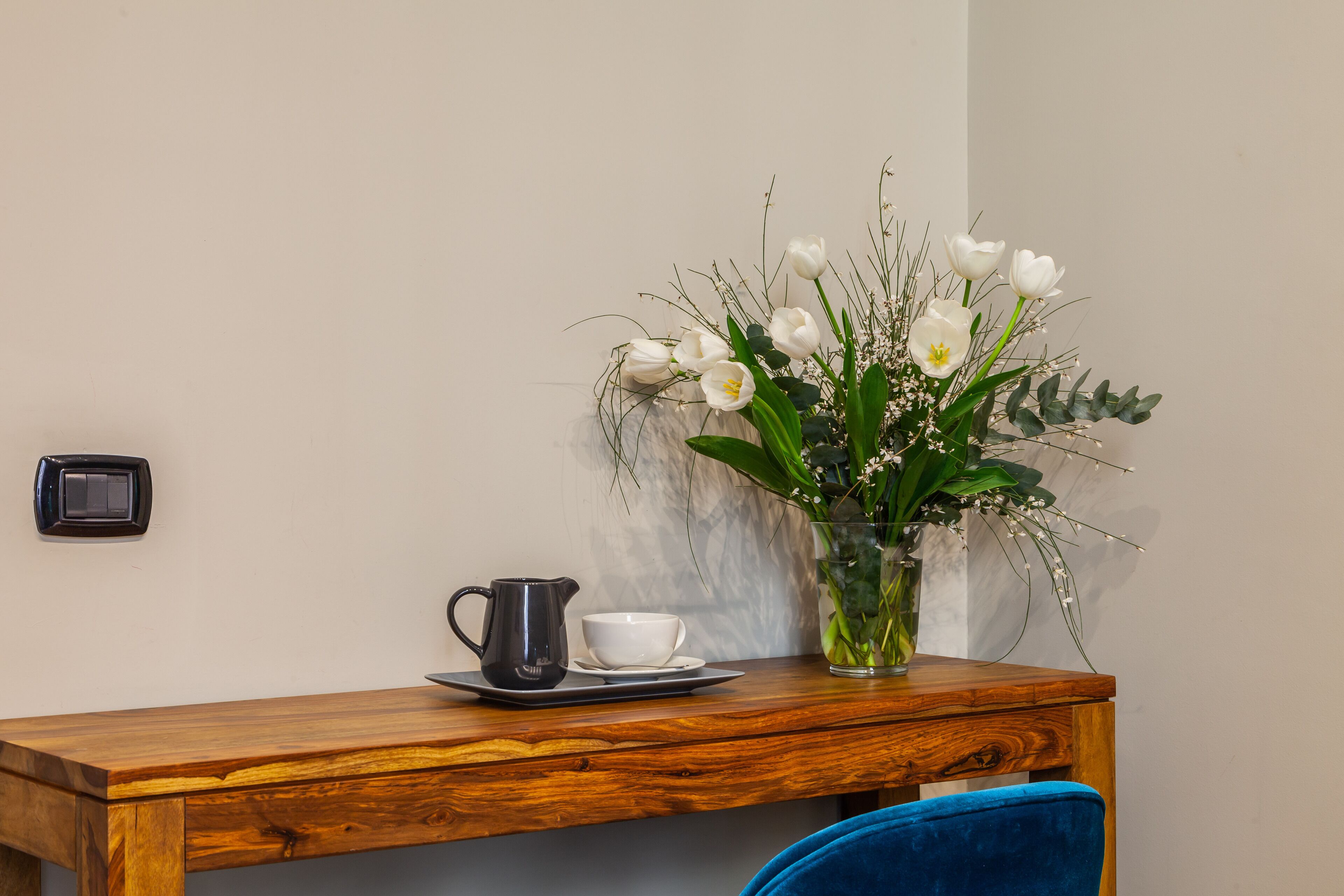 Een houten tafel met een zwarte kruik en een witte kop, een vaas met witte bloemen, en een blauwe stoel.