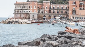 A historic building with arched windows and a castle-like facade, located near a rocky shoreline with boats and people. - Donna Lena Relais (Naples)