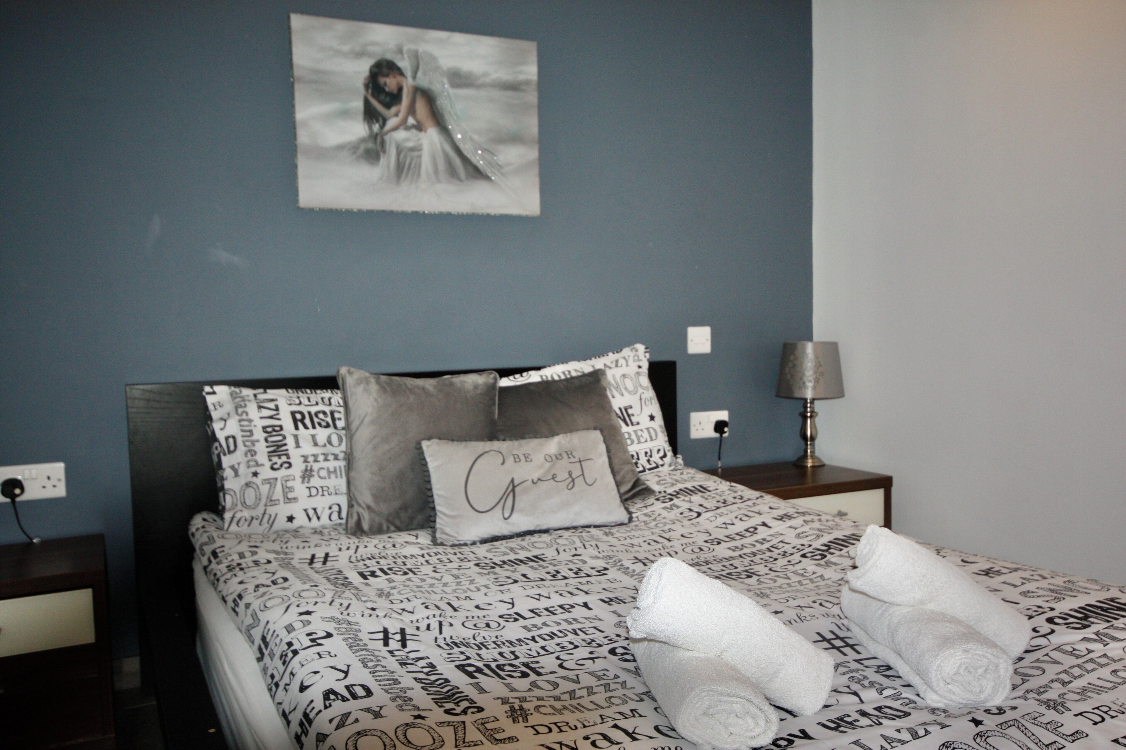A bedroom with a bed covered in a printed blanket, a pillow with the text "Be our guest," and a bedside lamp.