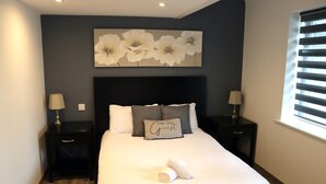 Ein ordentlich gemachtes Bett mit einem Kissen, auf dem "BE OUR GUEST" steht, und einem darüber gerollten Handtuch.