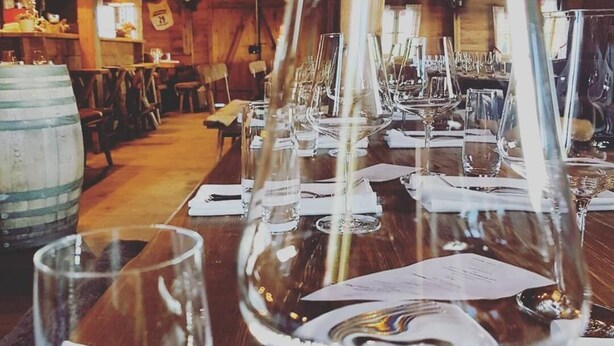 A long dining table set with wine glasses, plates, and cutlery, in a rustic wooden interior with exposed beams.