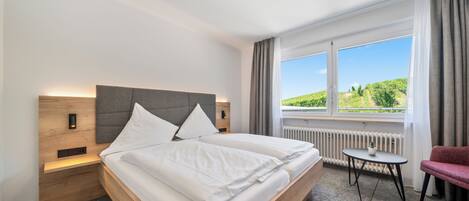 A modern hotel room with a large bed, a small table, a chair, and a view of greenery through the window.
