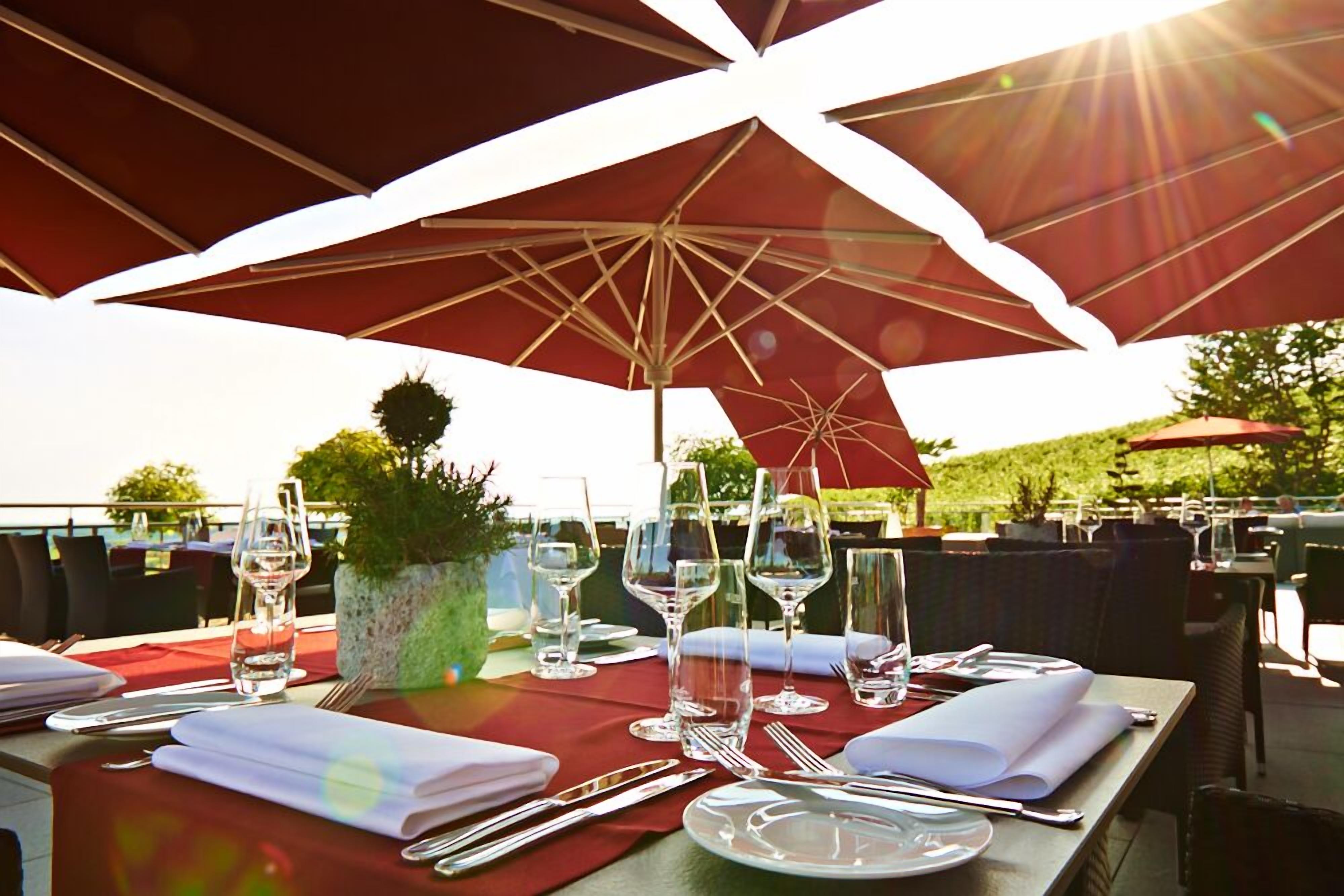 An outdoor dining area with tables set for two, under large red umbrellas, with wine glasses and cutlery on the table.