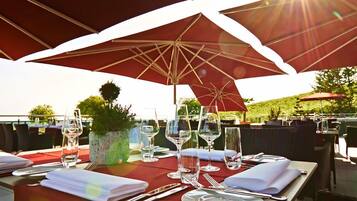 An outdoor dining area with tables set for two, under large red umbrellas, with wine glasses and cutlery on the table.
