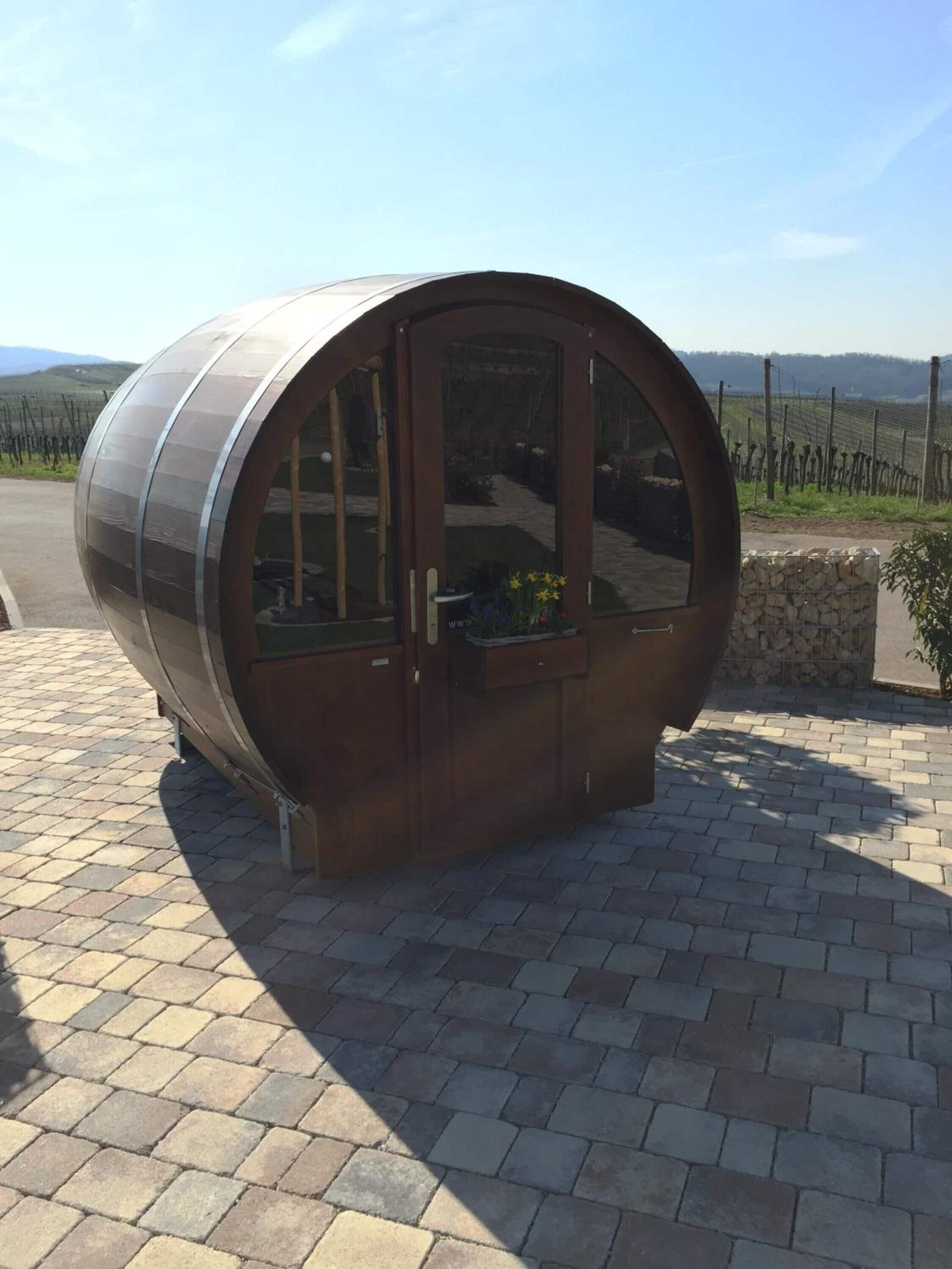 A cylindrical structure with a glass door, situated on a paved area with a stone wall and a vineyard in the background.