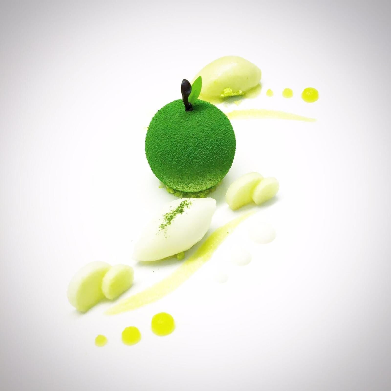 Una manzana verde con superficie rugosa, rodeada de pequeños puntos amarillos y un objeto blanco con motas verdes.