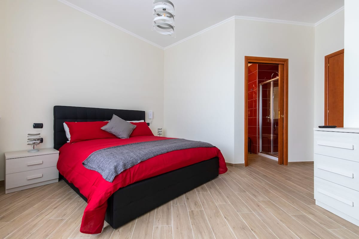Un dormitorio moderno con cabecera negra, ropa de cama rojo y gris, una mesita de noche blanca y suelo de madera.