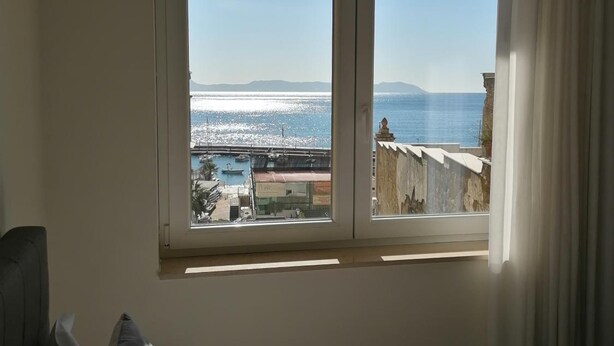 Un dormitorio con un ventanal que ofrece vistas al mar, con un puerto deportivo y montañas a la distancia.