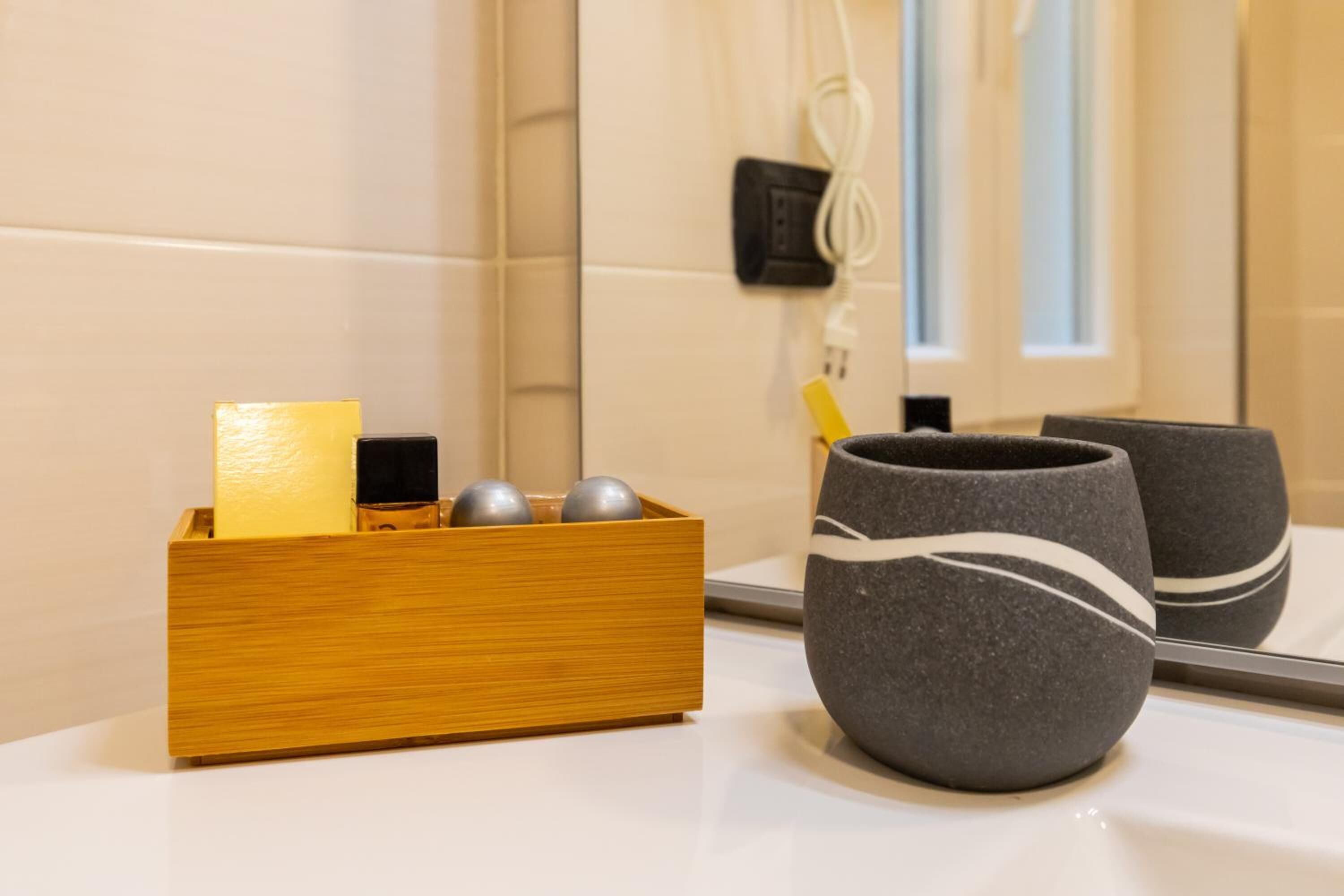 Une salle de bain avec un récipient en bois contenant un pain de savon et deux boules métalliques, ainsi que deux tasses en céramique foncée à rayures blanches, posées sur un comptoir blanc.