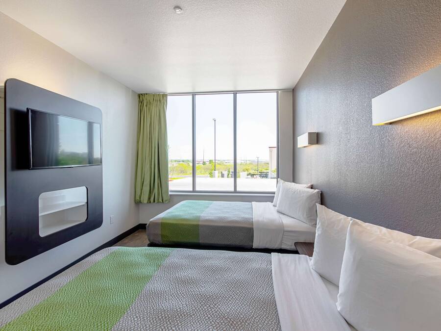 A hotel room with a large window, a flat-screen TV mounted on the wall, a bed with white bedding, and a green and white striped area rug.
