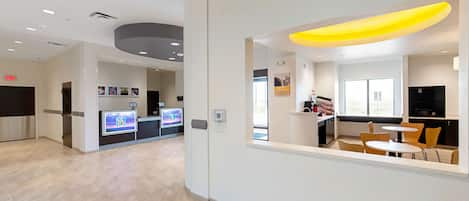Um lobby de hotel moderno com balcão de recepção, tela digital, área de estar e vista para o espaço de refeições.