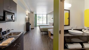 A hotel room with a kitchenette, a bed, a bathroom with a sink and mirror, and a view of the outdoors. - Studio 6 Austin, TX - Airport (Austin)