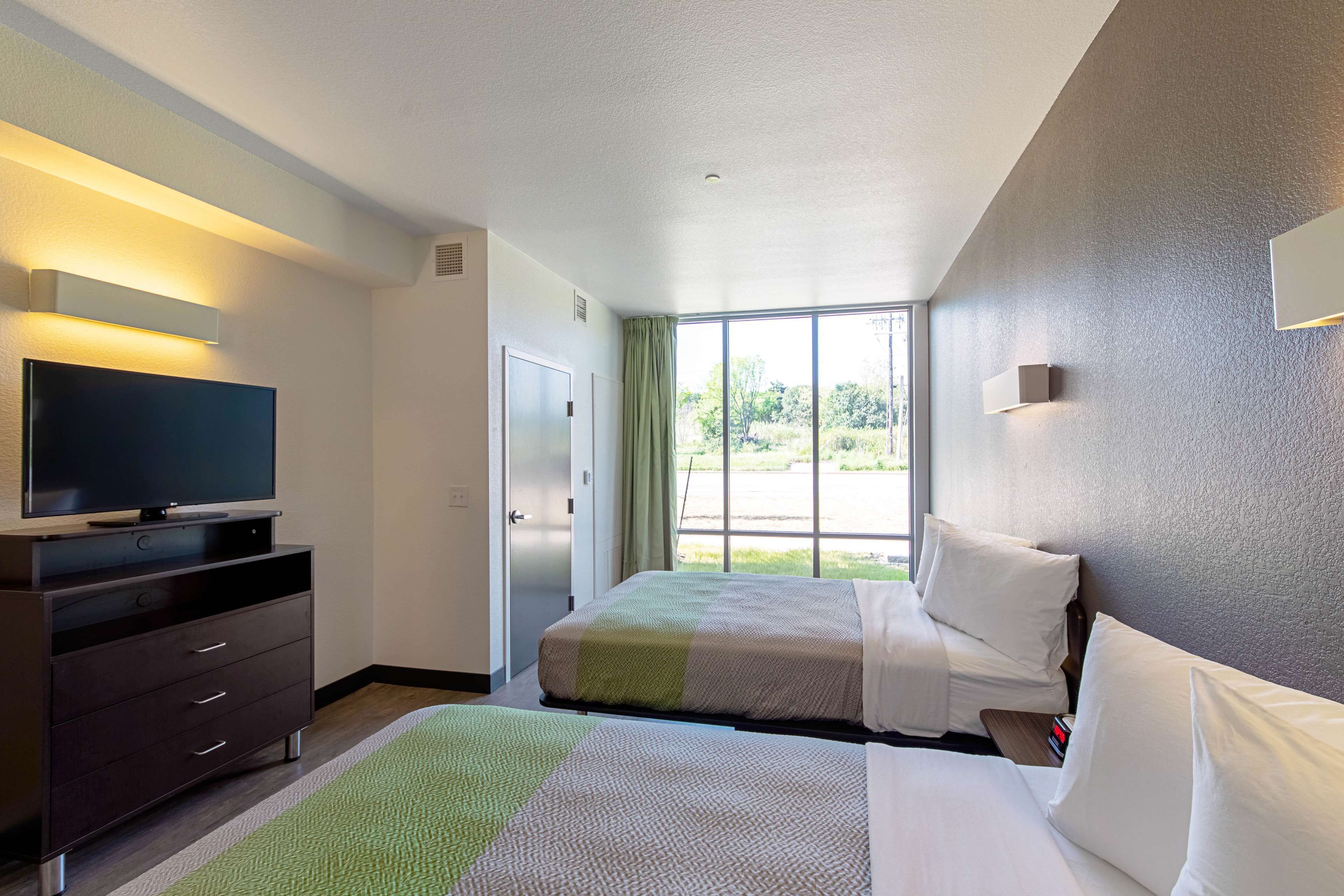 A hotel room with two beds, a TV, a dresser, and a large window offering a view of the outdoors.