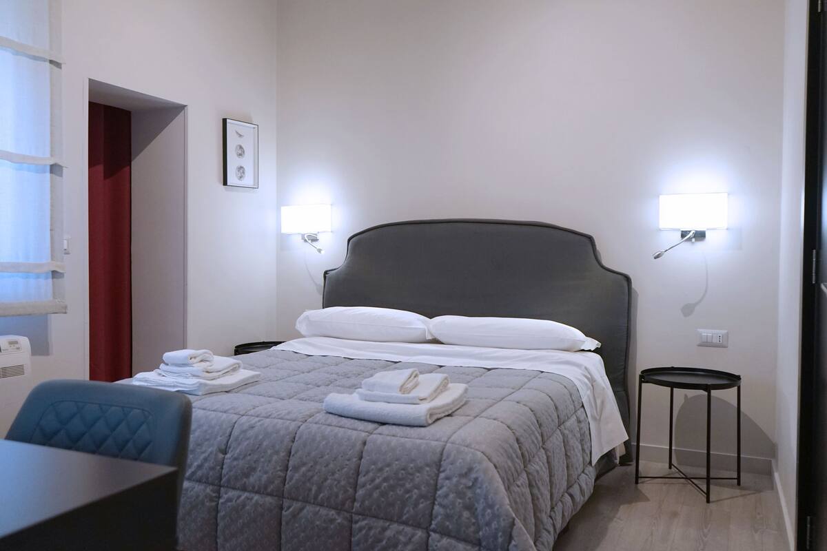 Une chambre d’hôtel avec un lit, un bureau et une chaise. Il y a deux lampes fixées au mur et un tableau accroché au mur.