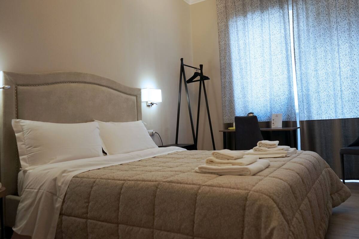 Un lit bien fait, recouvert d’une courtepointe beige, une table de chevet avec une lampe et une fenêtre avec des rideaux.