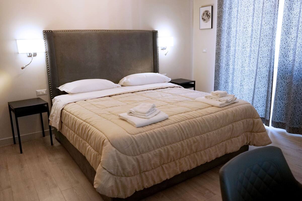 Un lit bien fait, recouvert d’une couverture beige, une table de chevet, une commode et une chaise.