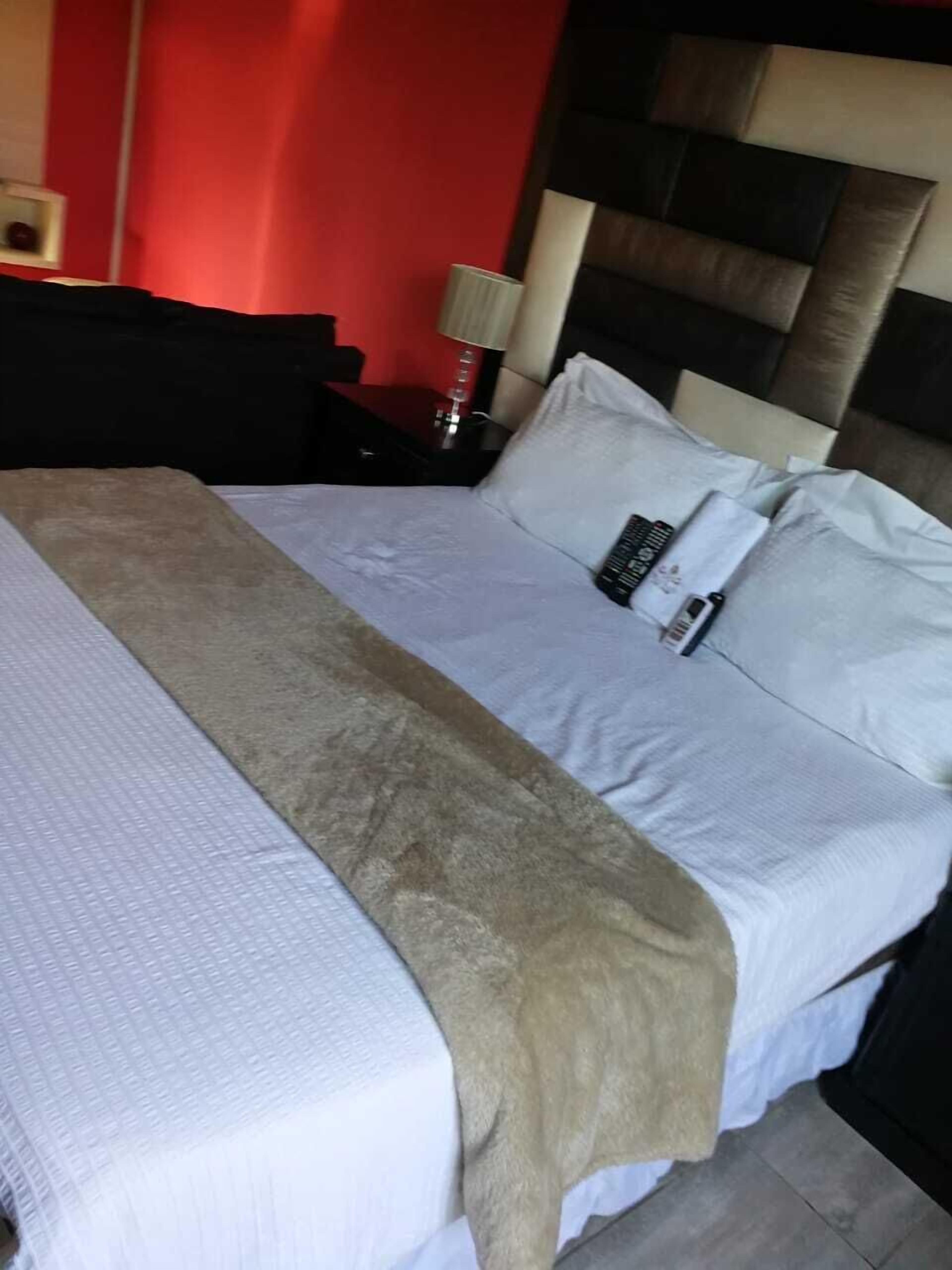 Una cama con una colcha blanca y beige, un control remoto y un cojín en una mesita de noche con una lámpara.