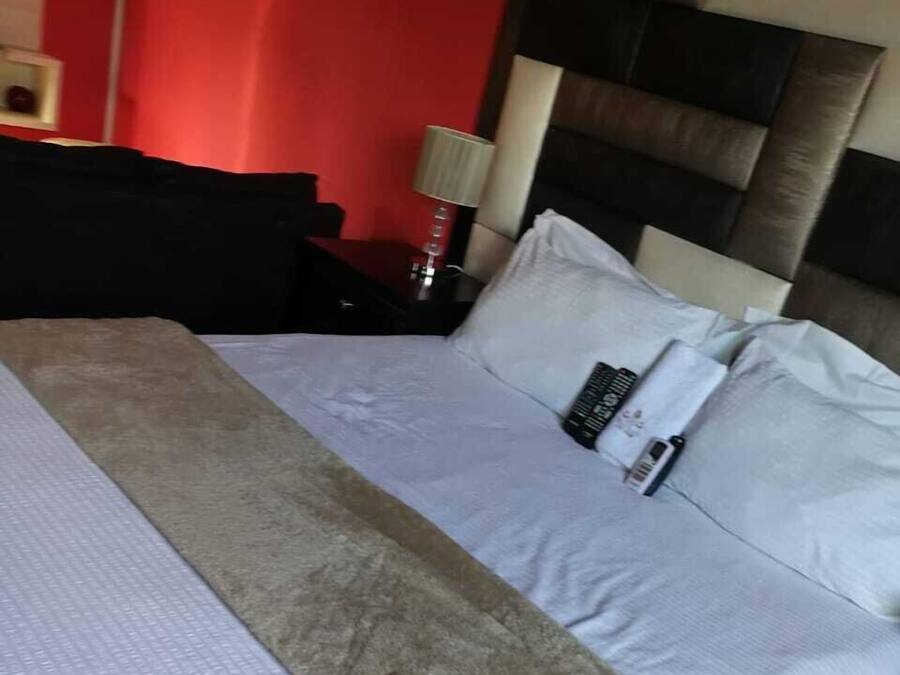 Una cama con una colcha blanca y beige, un control remoto y un cojín en una mesita de noche con una lámpara.