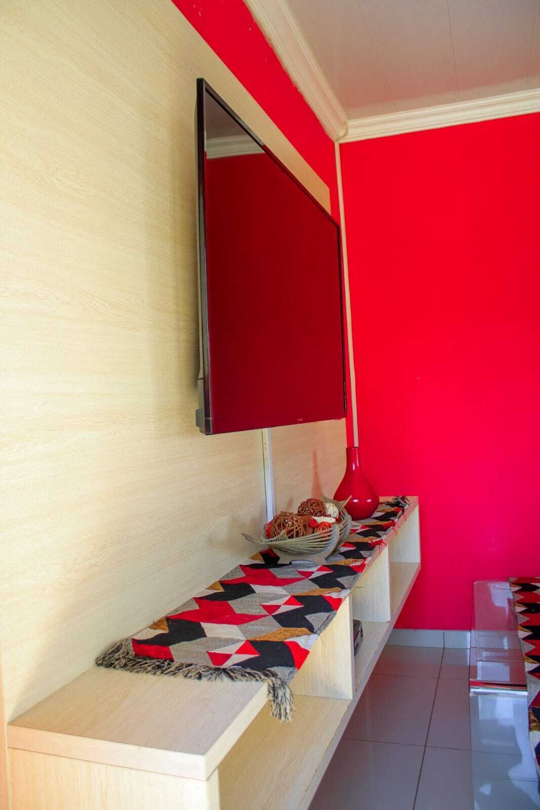 A room with a red wall, a mounted TV, a shelf with decorative items, and a patterned runner.