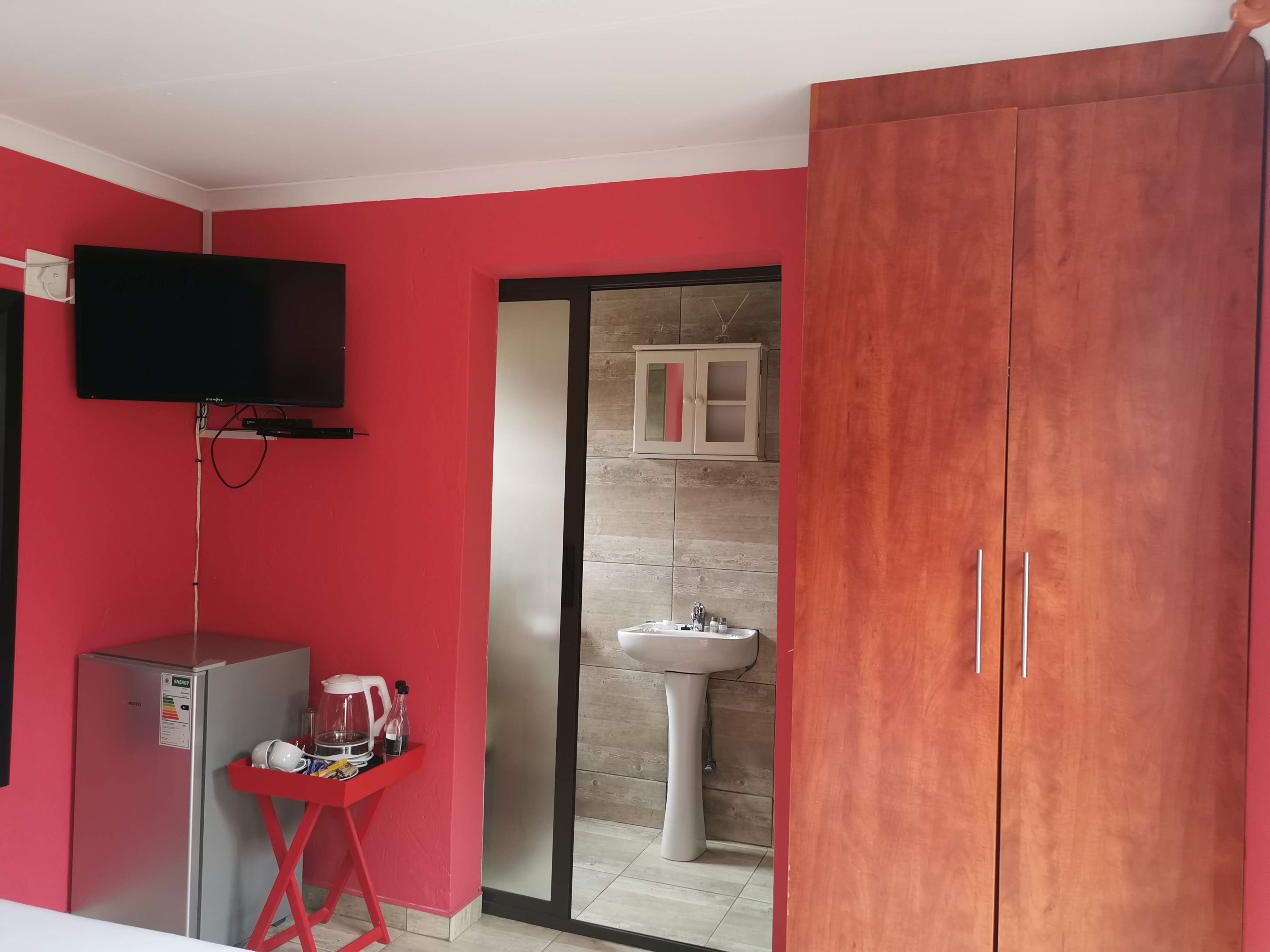 A room with red walls, a television mounted on the wall, a refrigerator, a small table with items, a wooden cabinet, and a sliding glass door leading to a bathroom with a sink and mirror.