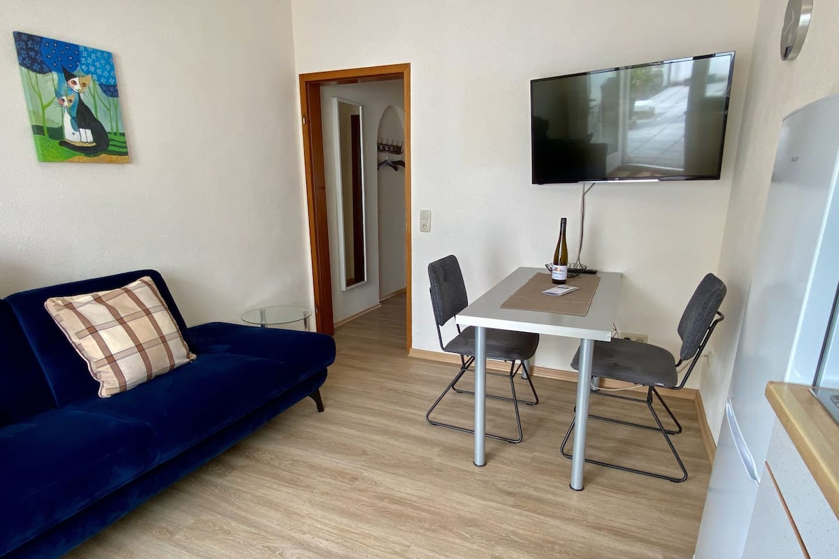 Uma sala de estar com um sofá azul, uma mesa de jantar com quatro cadeiras, uma televisão de tela plana na parede e um quadro na parede.