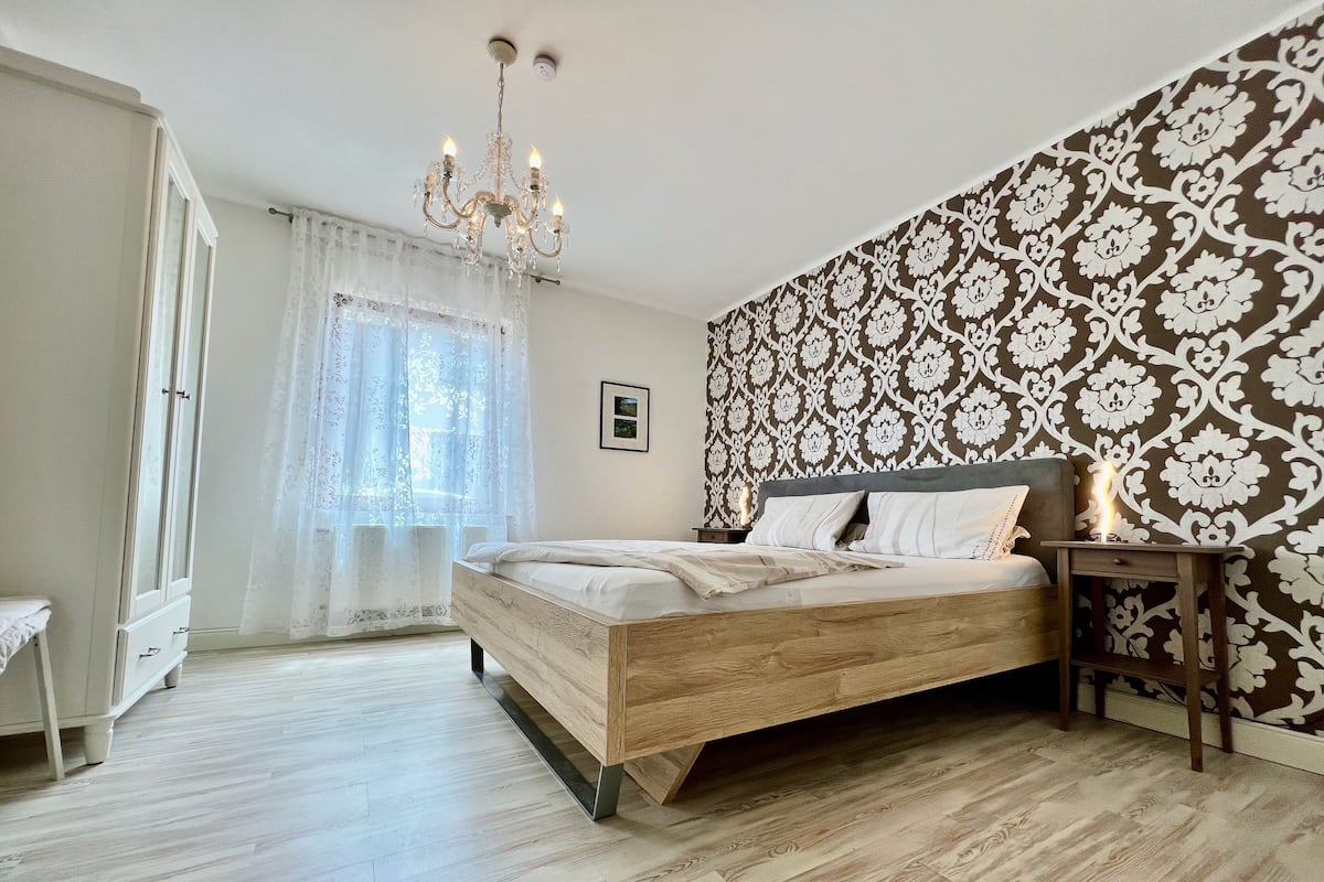 Quarto com cama de madeira, mesa de cabeceira, cômoda e parede com padrão.