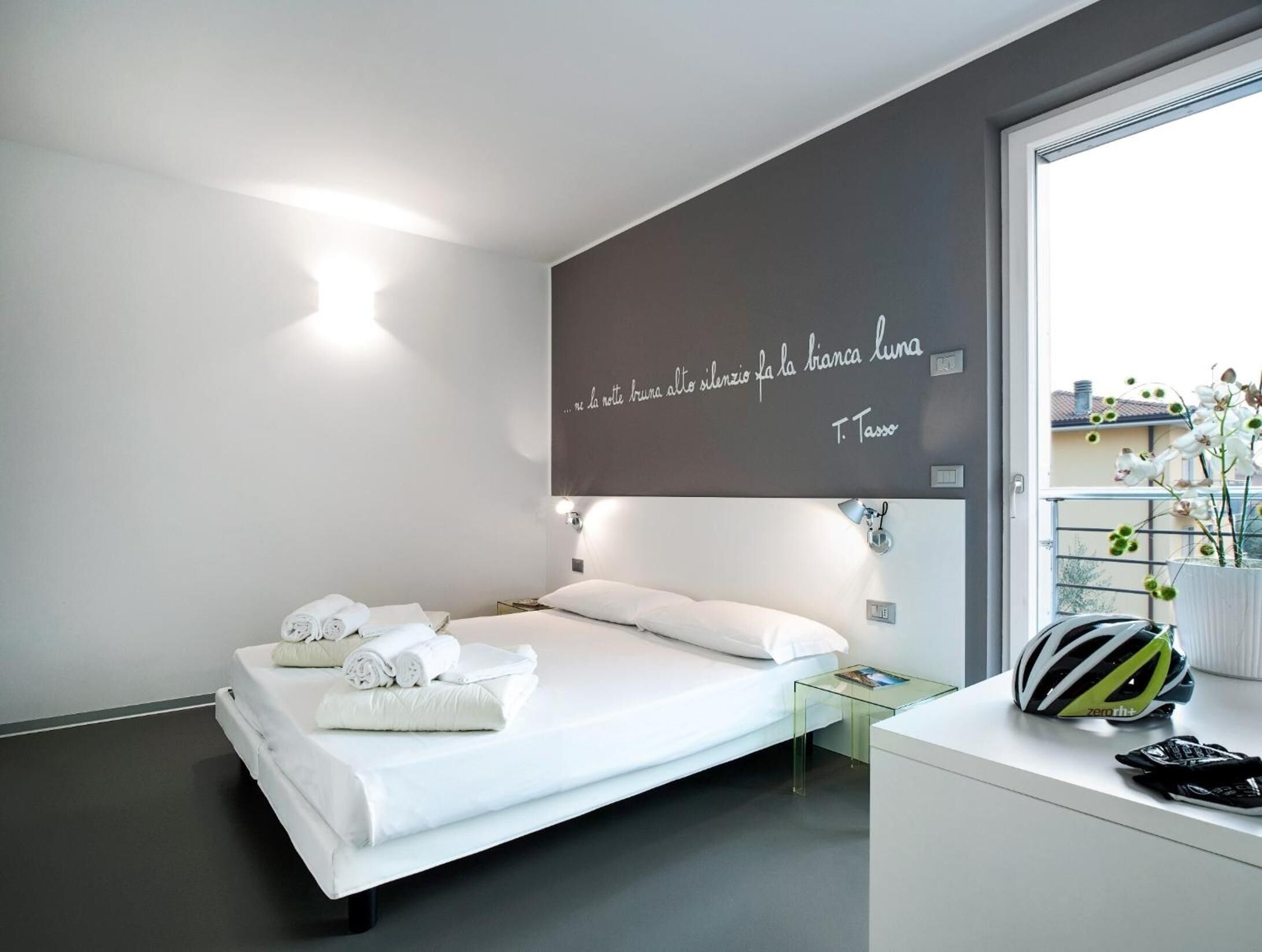 A modern hotel room with a bed, a glass table, a bicycle helmet, and a quote on the wall.