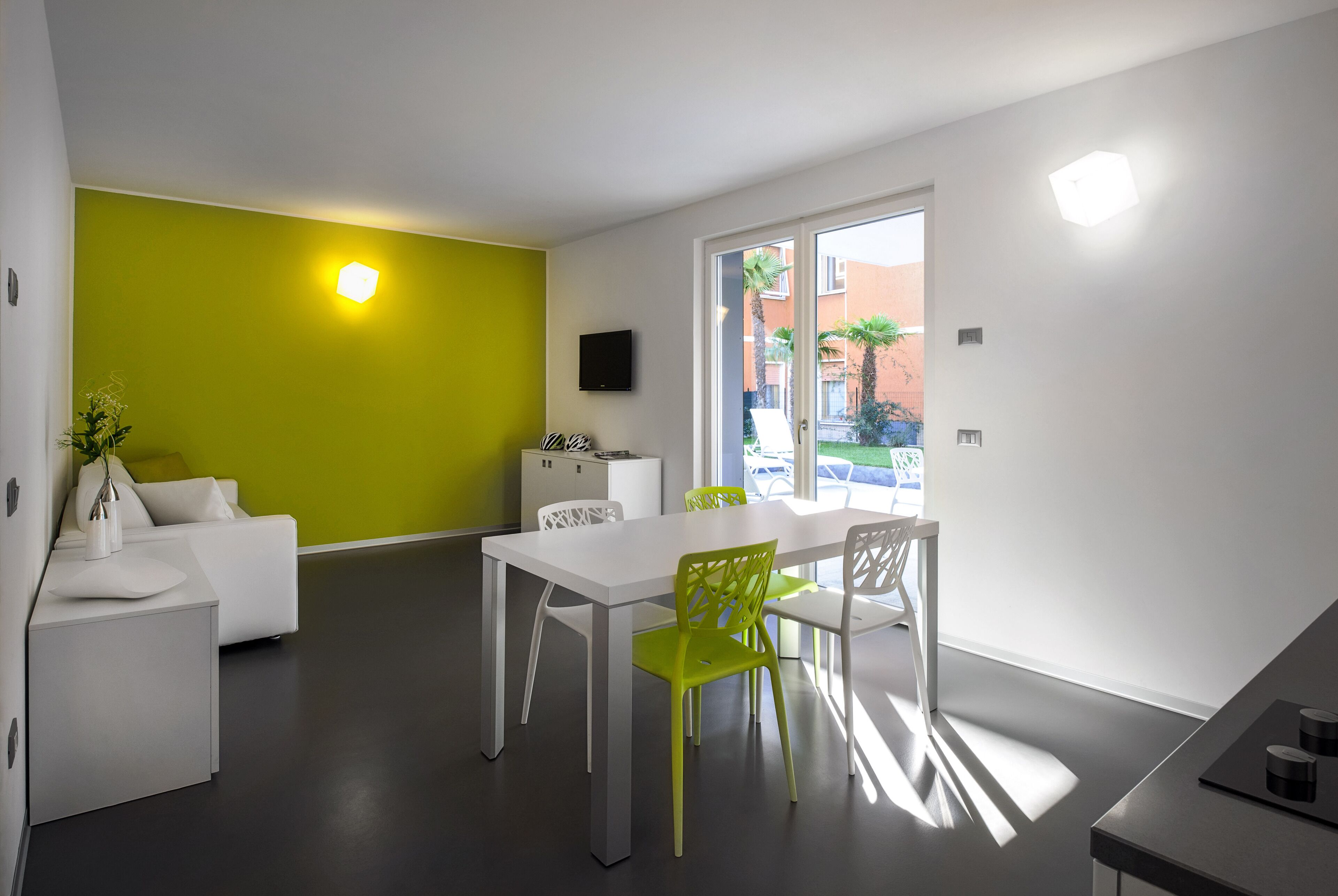 Sala moderna com sofá branco, mesa de jantar com cadeiras verdes, pequena área de cozinha e uma grande janela com vista para o exterior.