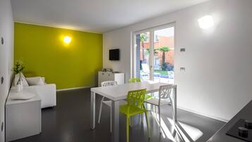 Sala moderna com sofá branco, mesa de jantar com cadeiras verdes, pequena área de cozinha e uma grande janela com vista para o exterior.