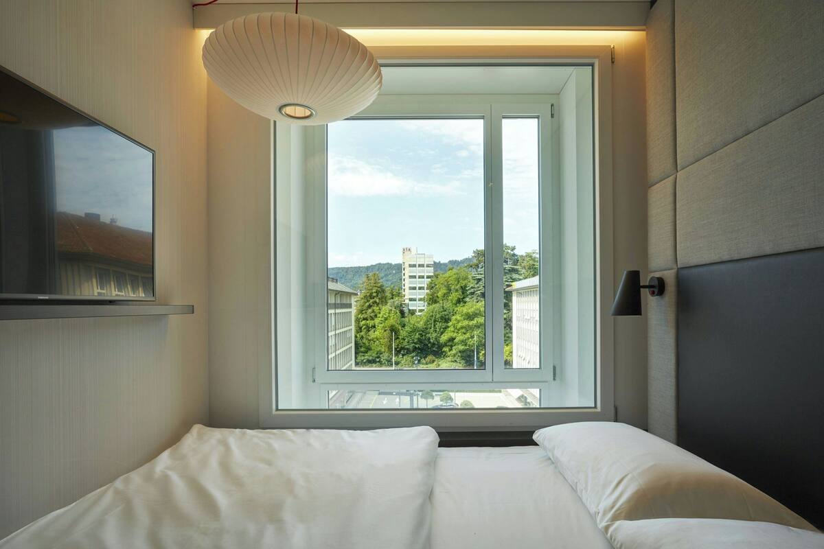 A modern hotel room with a large window, a bed, a flat-screen TV, and a wall-mounted lamp.