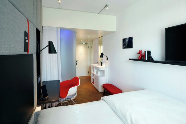 A modern hotel room with a bed, a desk with a chair, a vanity, and a TV on the wall.