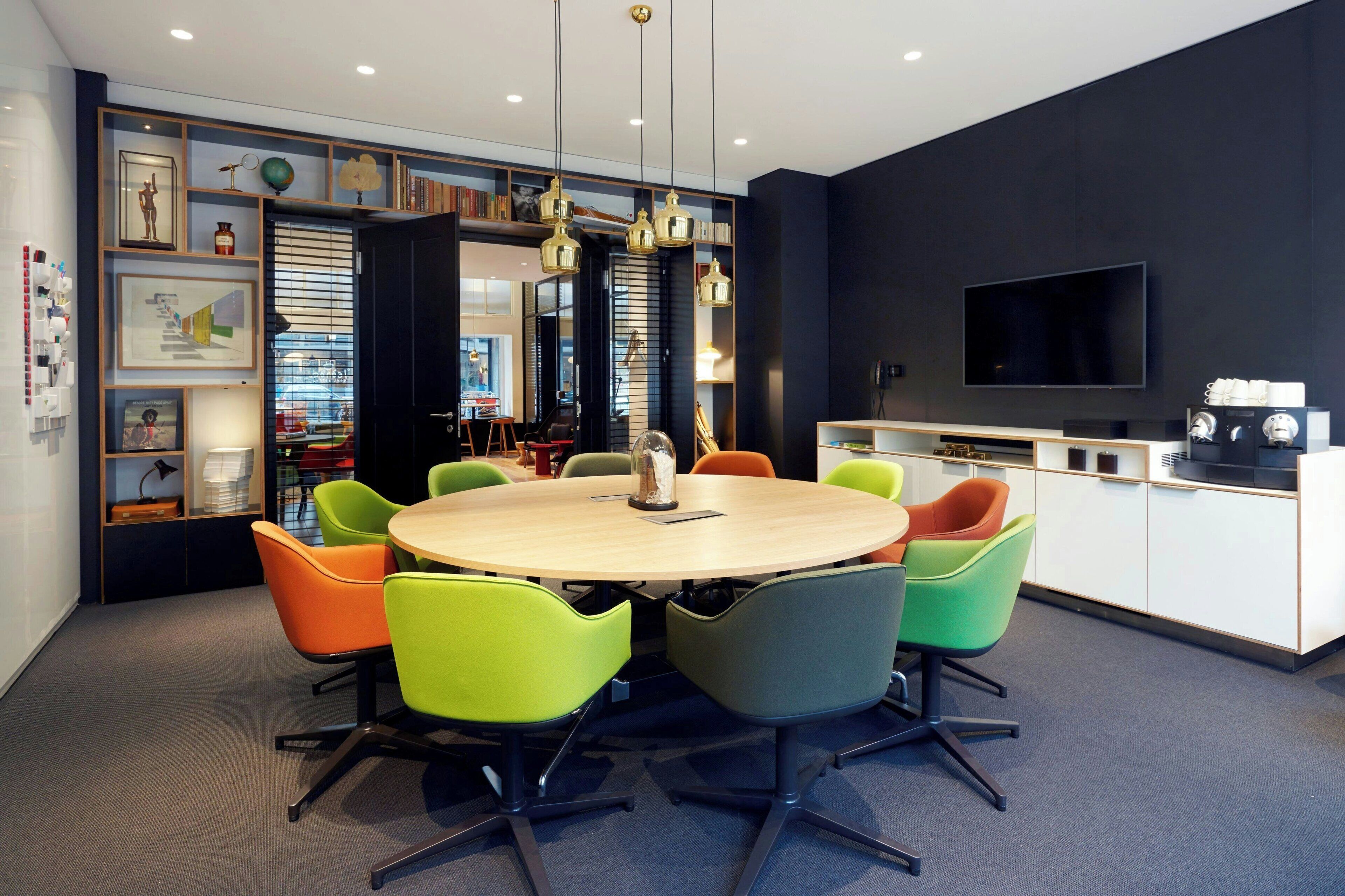 A modern dining area with a round table and colorful chairs, a kitchen with a TV, and a bookshelf with various items.