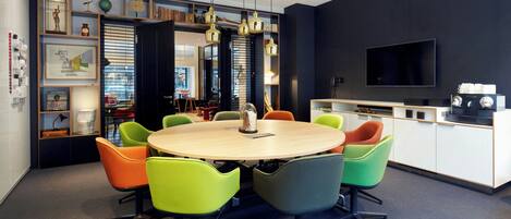 A modern dining area with a round table and colorful chairs, a kitchen with a TV, and a bookshelf with various items.
