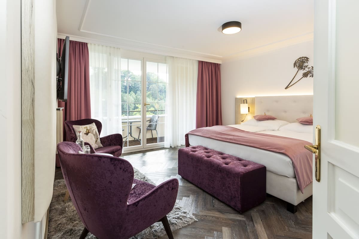 A hotel room with a large bed, a chair, a small table, and a view of the outdoors through a sliding door.