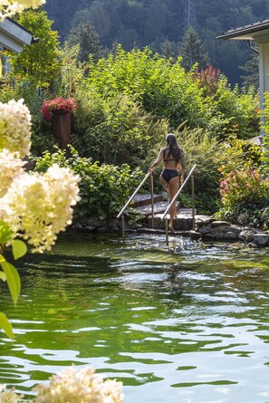 Une femme en bikini marchant sur un pont de bois qui enjambe un étang, le tout étant entouré d’une végétation luxuriante.