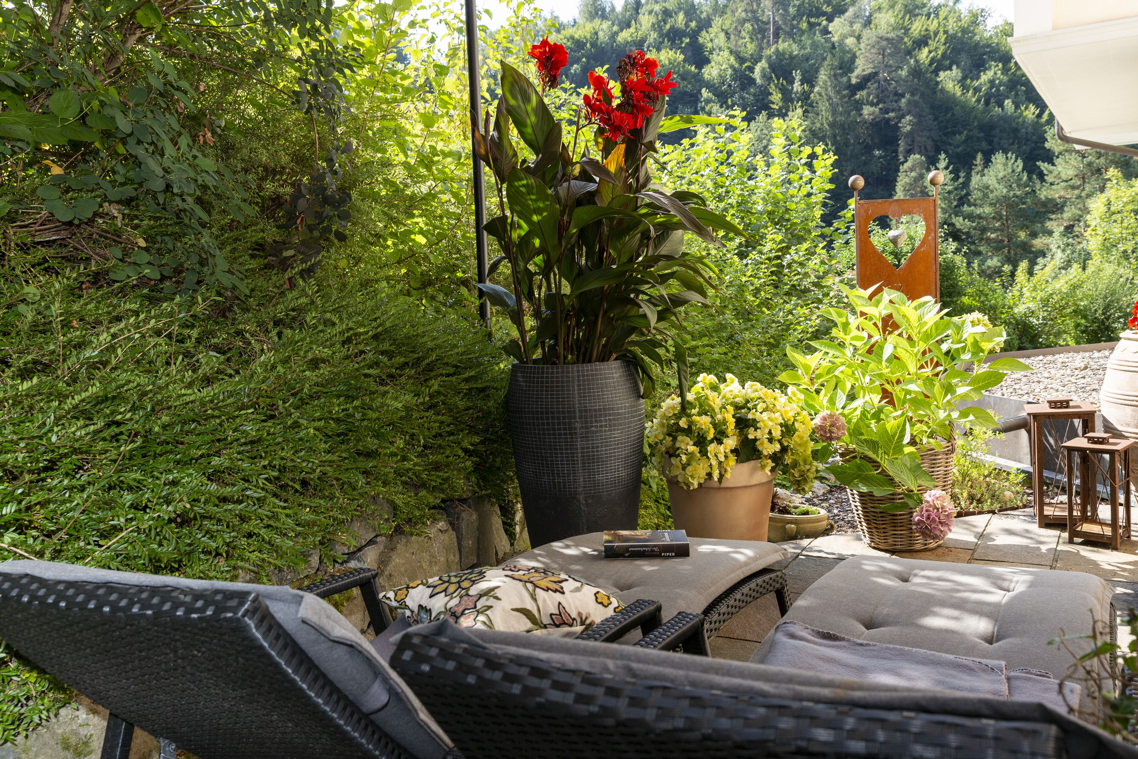 Un patio con mobiliario exterior, una planta en maceta con flores rojas y vista a un área boscosa.